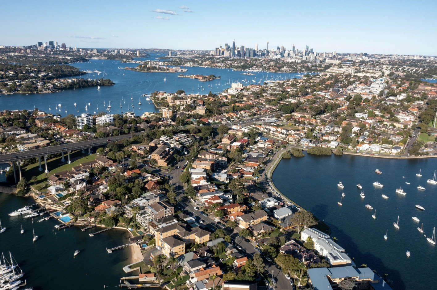 Aerial view of a harbor city with houses along the water, numerous boats, and a distant skyline on the horizon.